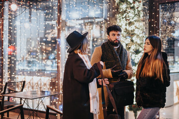 Three young adults engaged in a discussion outdoors at night surrounded by festive lights and urban backdrop.