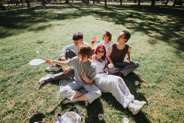 Five young adults sitting on the grass at a sunny park, sharing smiles and playing games, embodying friendship and leisure.