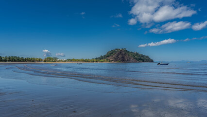 The waves of the ocean roll towards the shore, spreading along the beach. Hotel buildings, straw umbrellas in the distance. The boat is anchored.  A green hill against a clear blue sky. Copy space. 