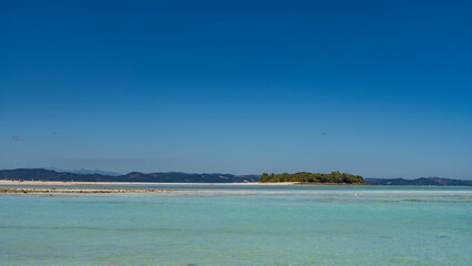 A tropical island covered with green vegetation and a sandbar are visible in the aquamarine ocean. Tiny silhouettes of people on a sandy beach. Mountains on the horizon. Clear blue sky. Copy space. 