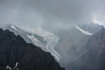 Dramatic alpine view to large glacier among rocks and ridges in gray low clouds in harsh weather....
