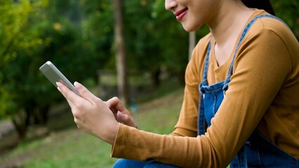 Asian woman using smartphone in orange orchard. Wearing denim overalls and brown long-sleeve shirt....