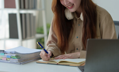 Young Asian woman looking happy with headphones around her neck Work on a laptop computer.online business Communicate with video conferencing Take notes in a notebook Ideas at the work desk.