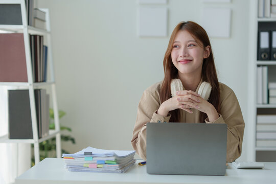 Young Asian woman looking happy with headphones around her neck Work on a laptop computer.online business Communicate with video conferencing Take notes in a notebook Ideas at the work desk.