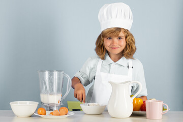 Portrait of a 7, 8 years old child in cook cap and apron making fruit salad and cooking food in kitchen. Cute little blonde happy smiling chef.