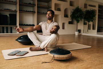 Serene portrait of calm African American young man in trance meditating in lotus pose at home...