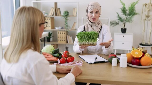 Mature lady sitting on appointment with muslim female nutritionist making healthy eating plan. Beautiful female doctor holding microgreen while adding it to patients diet.