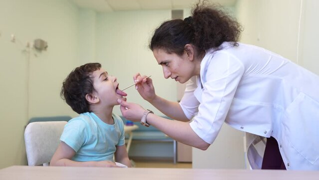 female doctor in a white coat examines a young boy's throat using a tongue depressor in a brightly lit clinic. The boy sits on a chair, cooperating with the check-up