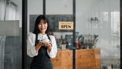 A woman wearing a black apron stands in front of a coffee shop with a sign that says "Welcome Open". She is holding a cup in her hand