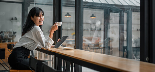 A woman is sitting at a table in a cafe, smiling and holding a cup of coffee. She is also holding a tablet and a laptop, which suggests that she might be working or browsing the internet