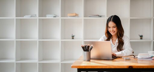 A woman is sitting at a desk with a laptop and a pencil holder. She is smiling and she is enjoying her work. The room is filled with shelves, and there are several books scattered around