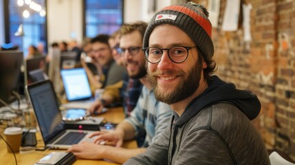 Smiling young professional wearing beanie hat and glasses sits at desk with coworkers in the background.