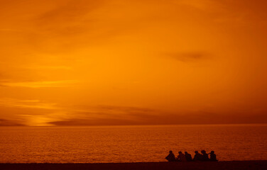 Young teenage people - guys and girls - sit on the beach, drinking beverages and watch the sunset. Group of five teen people sits on background. sunset beach. Travel or sea vacations. back, rear view.