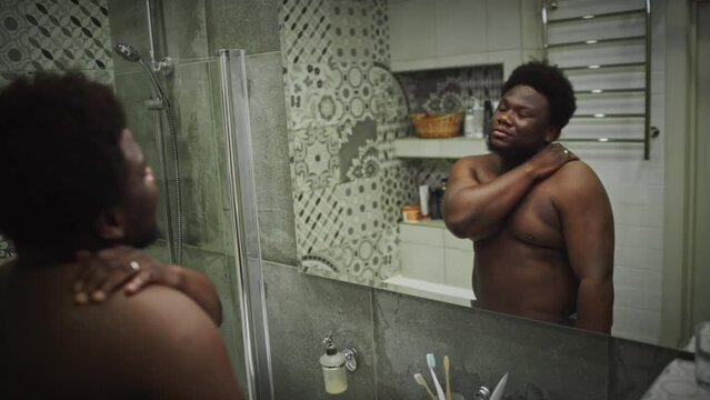 Young African American obese man with shirtless torso walking into bathroom at home and looking at his reflection in mirror