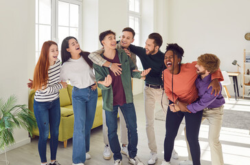 Portrait of a group of laughing happy young friends students or colleagues having fun hanging together at home. Guys and girls standing in a row, hugging and enjoying meeting. Friendship concept.