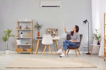 Young satisfied woman at home adjusts air conditioning in room using remote control. Woman sitting...