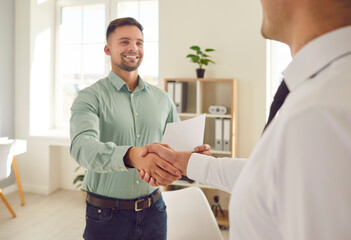 Obraz premium Two happy young men shaking hands and smiling. Two people meet in the office, establish partnership relations, make a lucrative business deal and exchange handshakes. Cooperation, teamwork concept