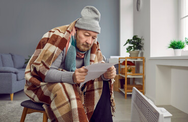 Senior man sitting at home wrapped in a blanket in winter hat and looking at the utility bill. A pensioner using a modern electric heater in winter in a cold room warming up. Heating problems concept.