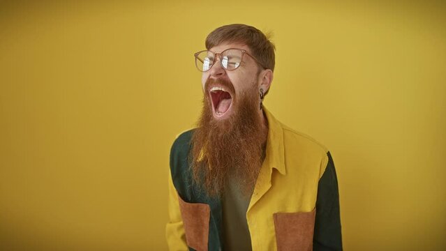 Frustrated young redhead man in glasses screams in fury, enraged and furious, showcasing aggression and rage over a yellow isolated background