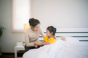 Young Asian mother sitting on bed looking at her daughter with smile of kind and happiness while...