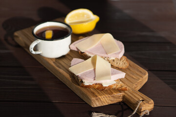 Sandwich with sliced ham butter and cheese on wooden cutting board near mug of tea with lemon