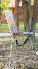 An artist's easel stands in a serene park setting, equipped with a wooden canvas, palette, and paintbrushes, capturing the beauty of nature under natural daylight