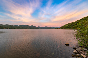 A serene sunset over a tranquil lake with rippled water, mountains in the distance, and a clear sky with soft clouds...
