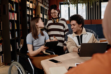 Caucasian girl with disability and her Asian and Middle Eastern friends studying in school library using digital tablet and laptop