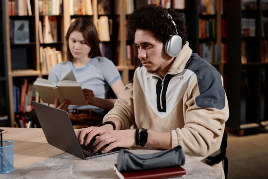 Medium shot of Middle Eastern male student spending time at university library typing on laptop, Caucasian girl reading book in background
