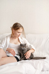 Teenager girl sitting on bed together with gray cat, stroking and embracing her pet