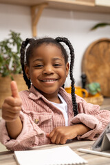 Black schoolgirl learning at home with textbook sitting at desk doing lessons showing like gesture