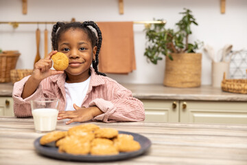 Cute little black girl sitting at table having snack milk and cookies in kitchen