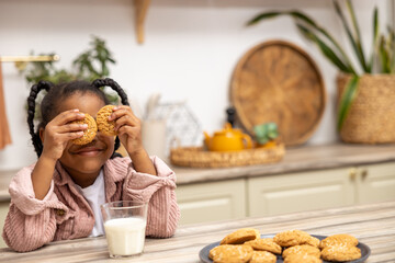 Little black girl sitting at table having snack covering eyes with cookies in kitchen