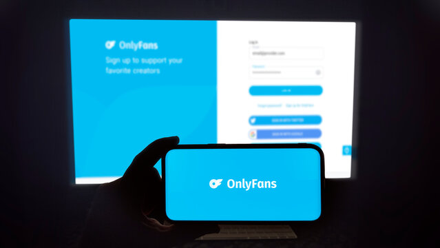 OnlyFans logo shown on a modern mobile phone. The smartphone is being held in landscape mode, in the darkness. A PC monitor with the OnlyFans website in the background.