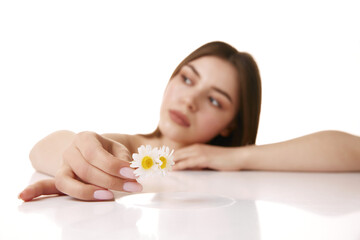 Young, beautiful woman with gentle expression, holding daisy looking away against white studio background. Selective focus. Concept of natural beauty and appearance, spa procedures and facial care.