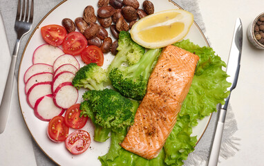Healthy lunch with grilled salmon on green lettuce, next to vegetables, tomatoes, radishes, broccoli and a portion of beans.