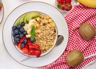Granola with strawberries, kiwi, banana and blueberries in a round plate on a white table.