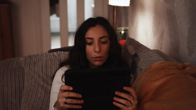 Young Italian woman smiles while relaxing using a tablet on the sofa