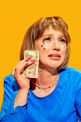 Portrait of young girl with makeup holding dollar money and crying against vivid yellow background....