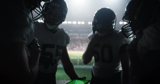 American Football Players Using Tablet Computer to Go Over Game Strategy Before Walking on a Stadium Filled with Energetic Sports Fans and Team Supporters. Multiethnic Footballers Getting Ready