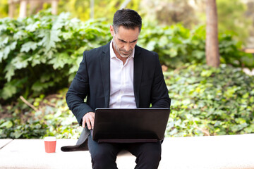 A middle-aged adult man in a suit, drinking coffee, looks at his laptop.