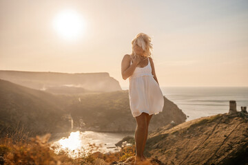 A blonde woman stands on a hill overlooking the ocean. She is wearing a white dress and she is...