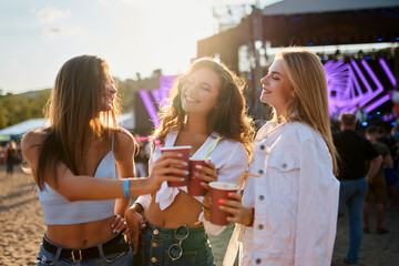 Group of women toast with cups at beachside music event. Smiling friends enjoy summer festival...