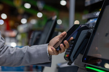 Image shows a person using a smartphone for NFC (Near Field Communication) mobile payment on a point-of-sale terminal in a retail environment. The focus is on the technology and convenience.