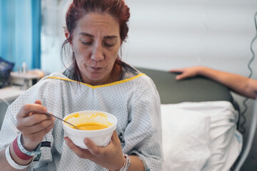 Redheaded woman on hospital bed, stirring puree with a spoon, looking sad and in pain during...