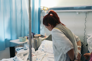 Young redheaded woman seen from behind adjusting hospital gown in hospital room post-surgery.