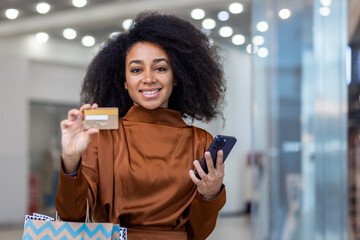 A cheerful woman with curly hair holding a credit card and smartphone, standing in a modern,...