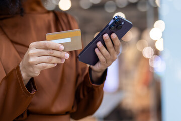 A person in a brown coat is shopping online, using a credit card and smartphone, in a shopping center with bokeh lights. They are making a payment and a purchase digitally with their mobile device