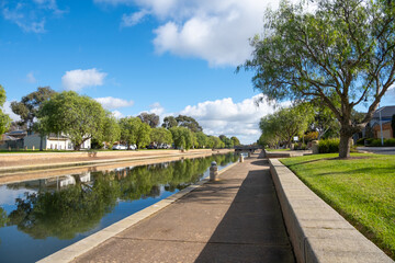A water canal is lined with trees and houses in a clean, beautiful residential neighborhood. A waterfront public footpath in Sanctuary Lakes, Point Cook, Melbourne Australia.