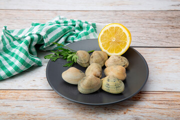 Raw clams on a black dish with a lemon half and a sprig of parsley, ready for cooking or seafood dishes
